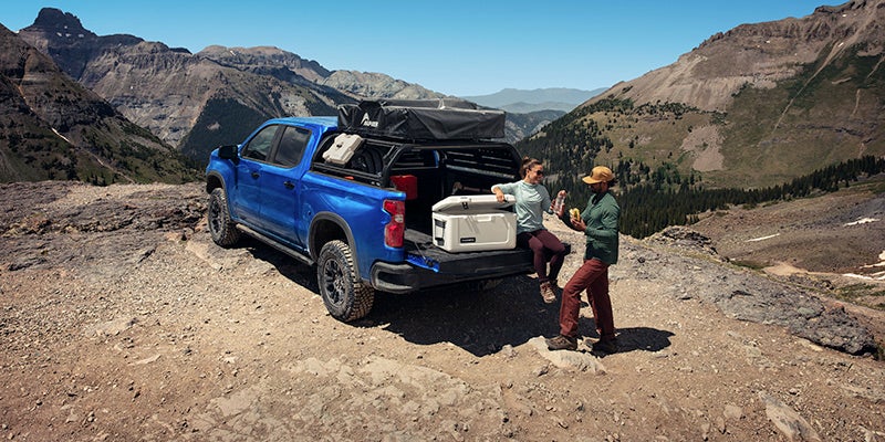 Blue Chevrolet Silverado 1500 parked with a mountain backdrop, couple relaxing in the truck.
